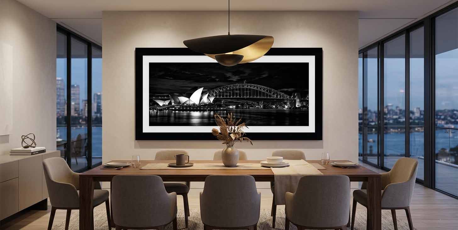 The Sydney Opera House and Harbour Bridge at night in black and white, framed in matte black with a white linen liner above a dining table in a modern room.