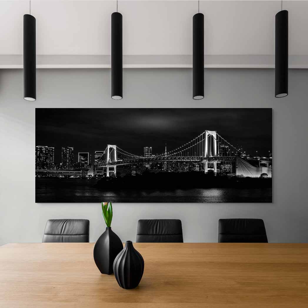 Wide black and white print of the Rainbow Bridge at night over Tokyo Bay, hanging on a light gray wall in a modern office conference room, above a wooden meeting table with black leather chairs and two dark vases, lit by four black cylindrical pendant lights overhead.