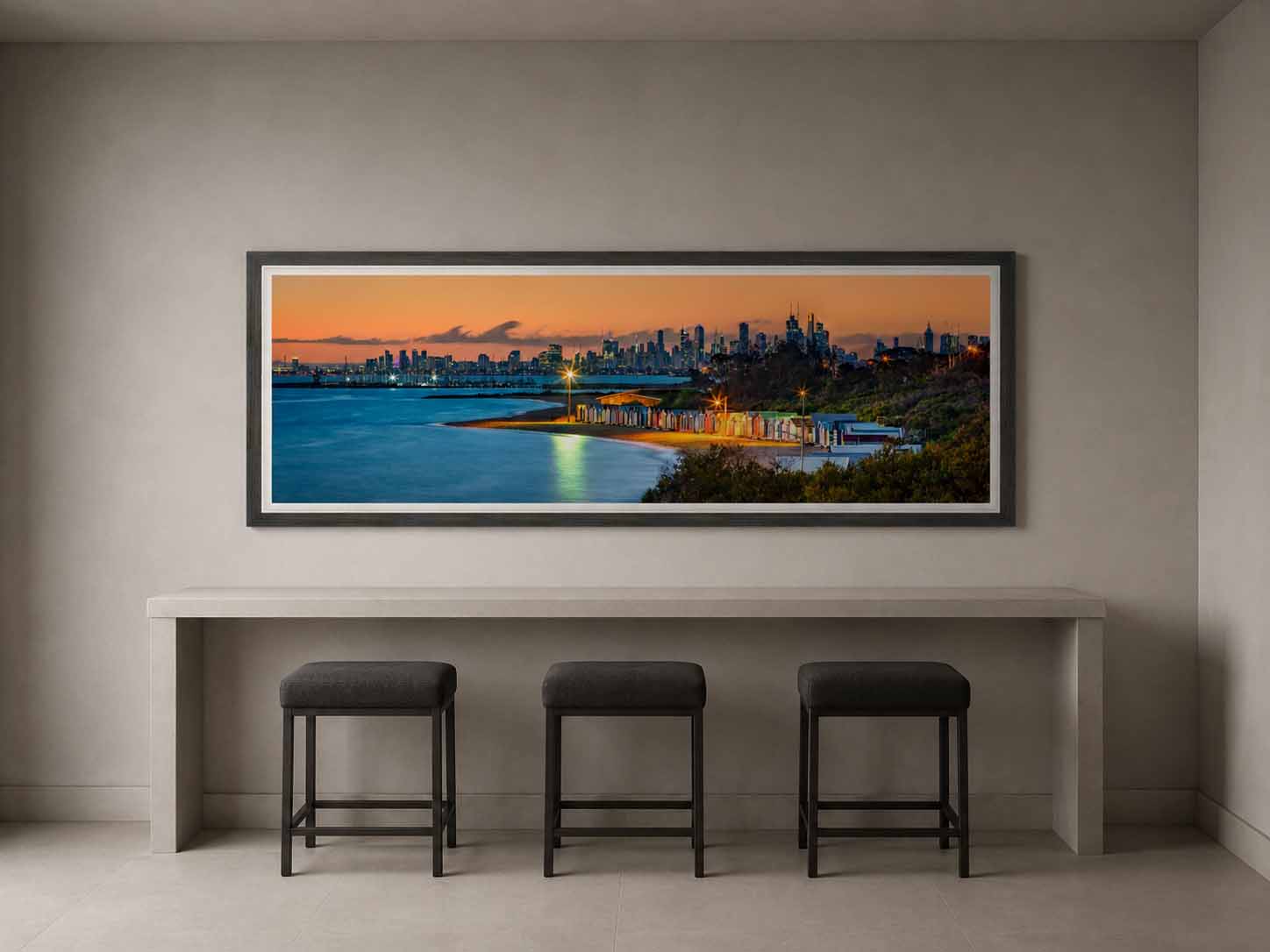 The Brighton bathing boxes at dusk with the Melbourne skyline beyond, framed in gray with a white linen liner above a stone counter and three stools.