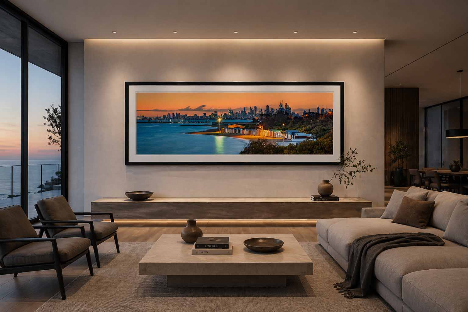 A large framed panoramic print of a lit beach at dusk with a city skyline hanging above a stone console in a modern coastal living room, with ocean visible through the windows