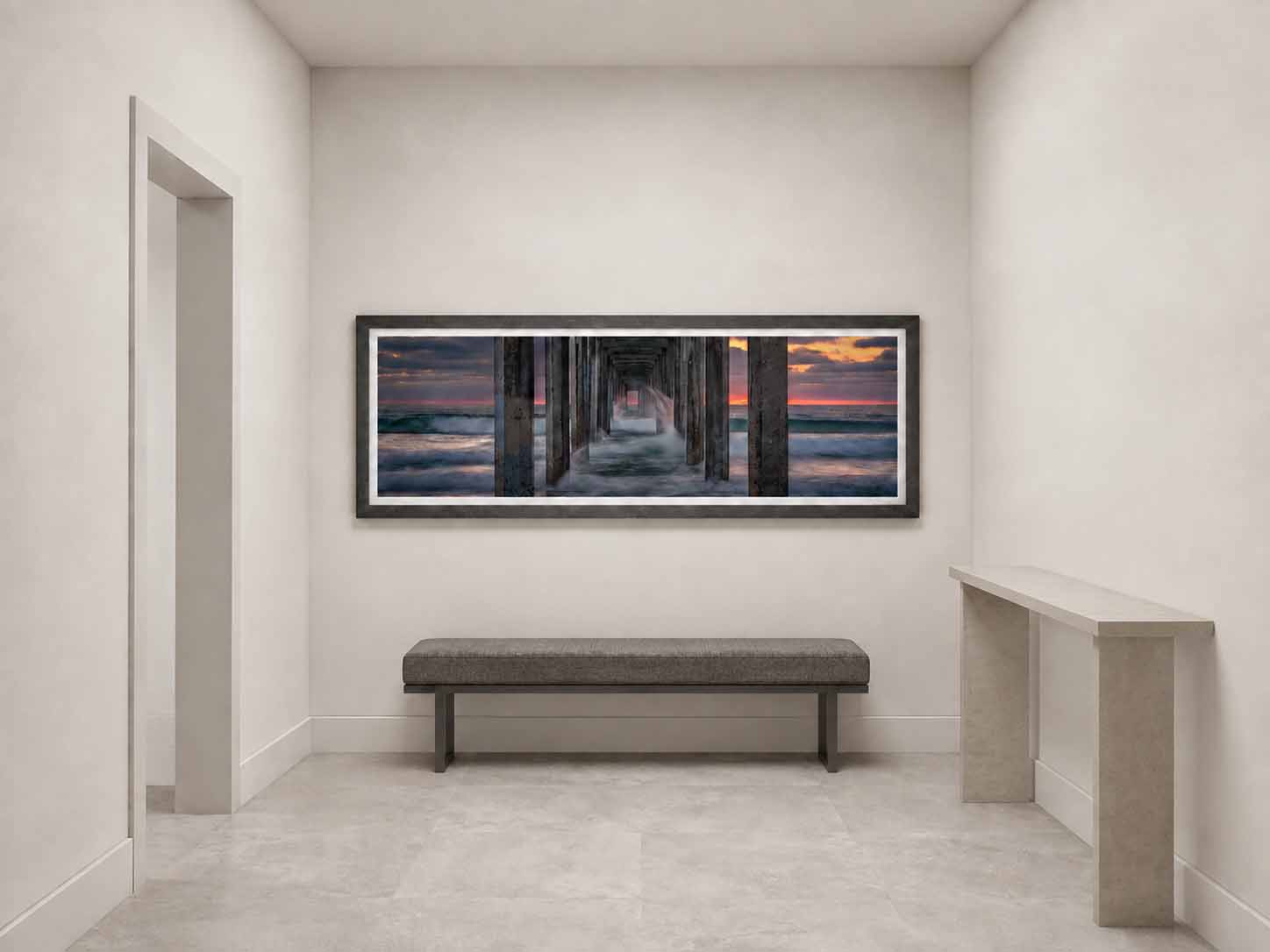 Waves breaking against the pilings beneath Scripps Pier at sunset, framed in matte black with a white linen liner on a pale wall at the end of a hallway.