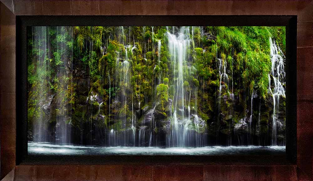 A photograph of a mossy waterfall in a dark ROMA frame with a black linen liner, hanging on a textured wall.
