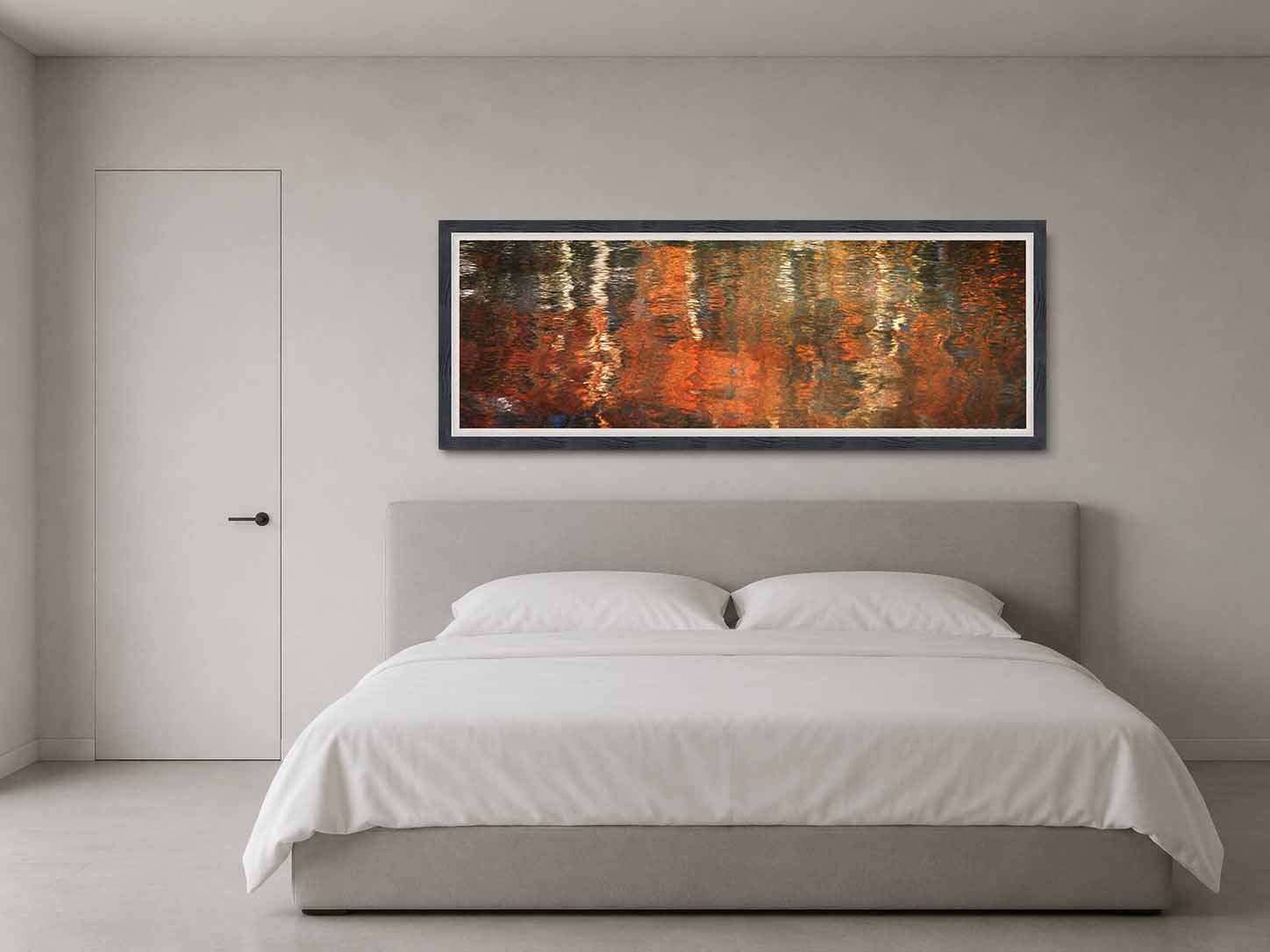 Autumn color reflected in moving water, framed in near-black with a white linen liner above a bed with white linens in a quiet gray bedroom.