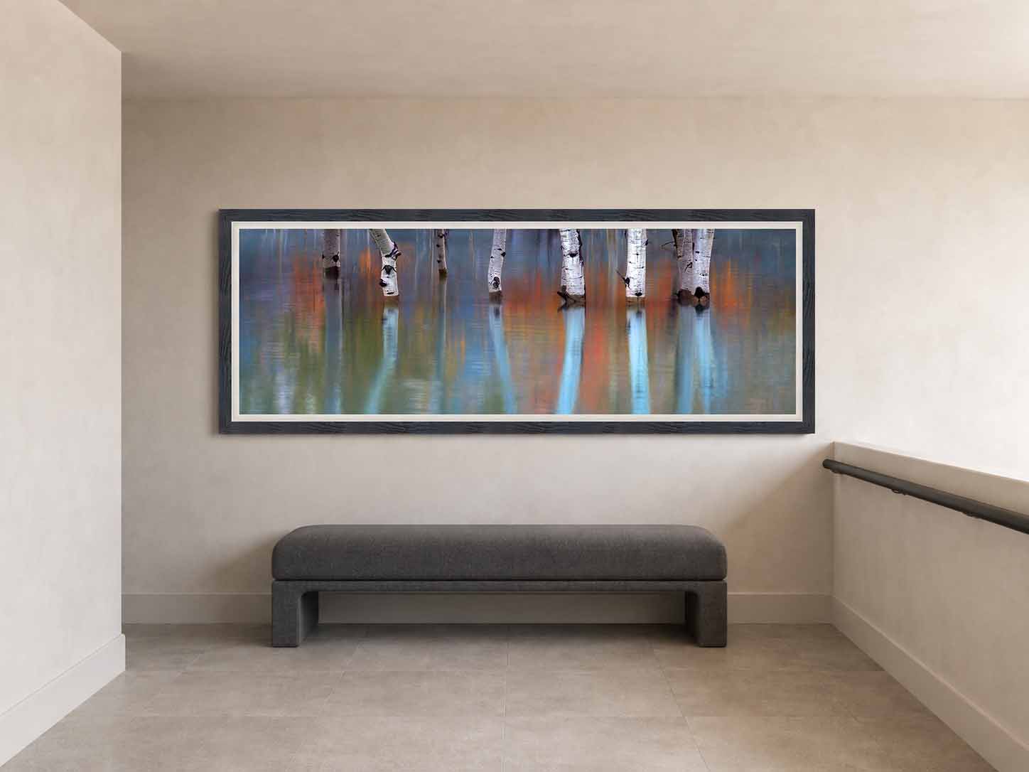 Aspen trunks standing in still water with autumn color reflected around them, framed in dark wood with a white linen liner on a pale wall at a stair landing.