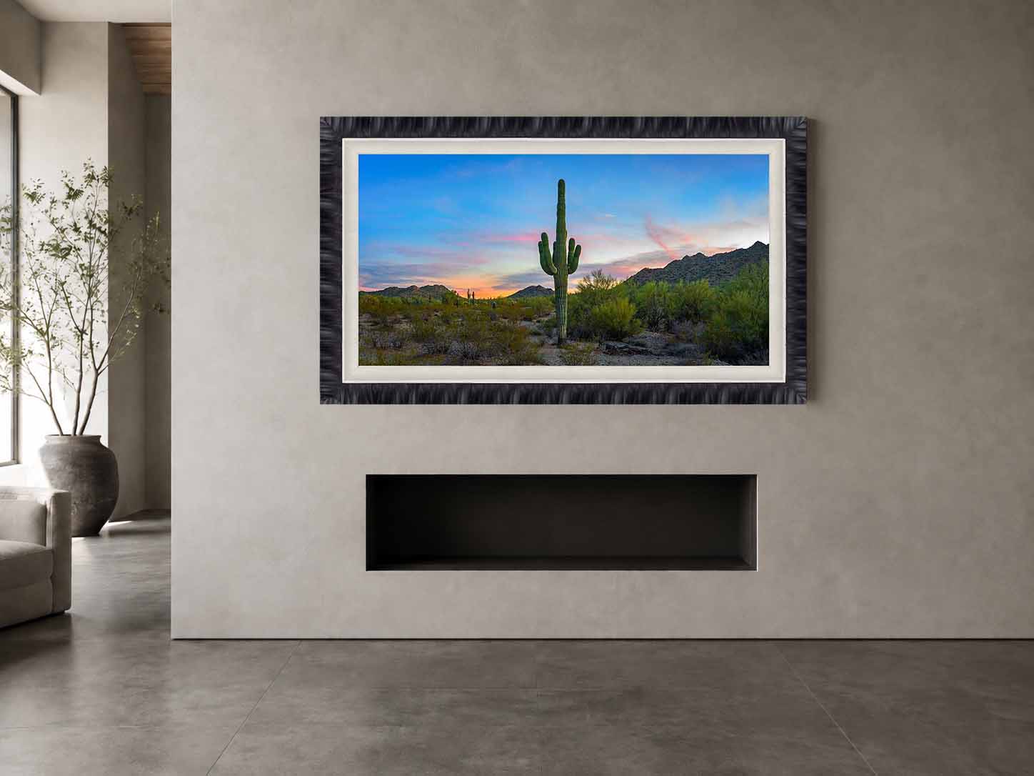 A photograph of a saguaro at twilight in the Sonoran Desert, framed in textured charcoal with a white linen liner, above a linear fireplace in a plaster wall.