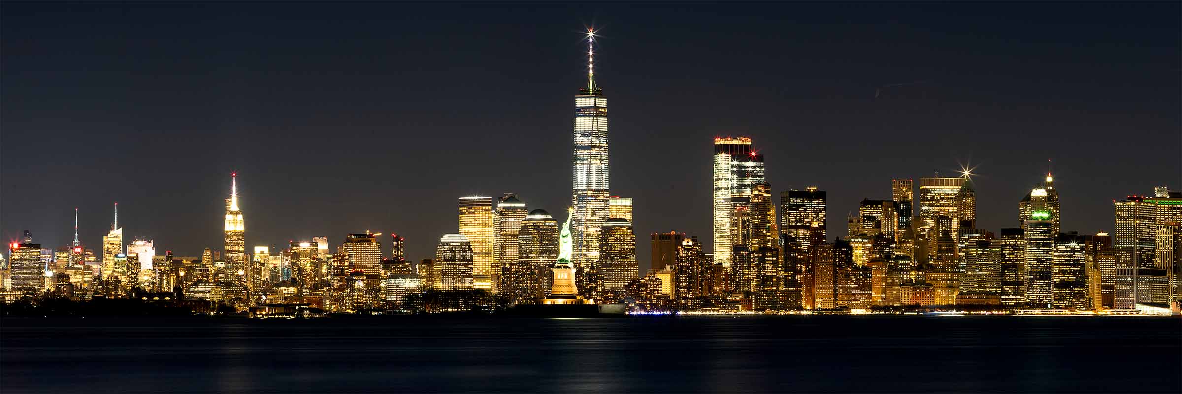 Statue of Freedom Night view of the New York City skyline with One World Trade Center illuminated, the Statue of Liberty centered, and dark water in the foreground.