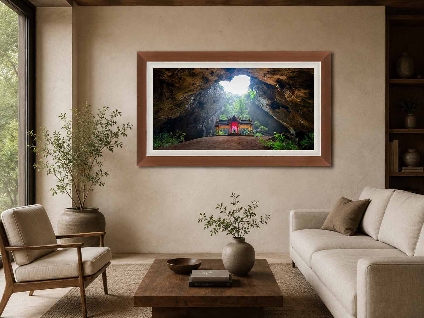 A photograph of a royal pavilion lit through a collapsed cave roof in Thailand, framed in dark walnut with a white linen liner, above a low table in a plaster-walled living room.
