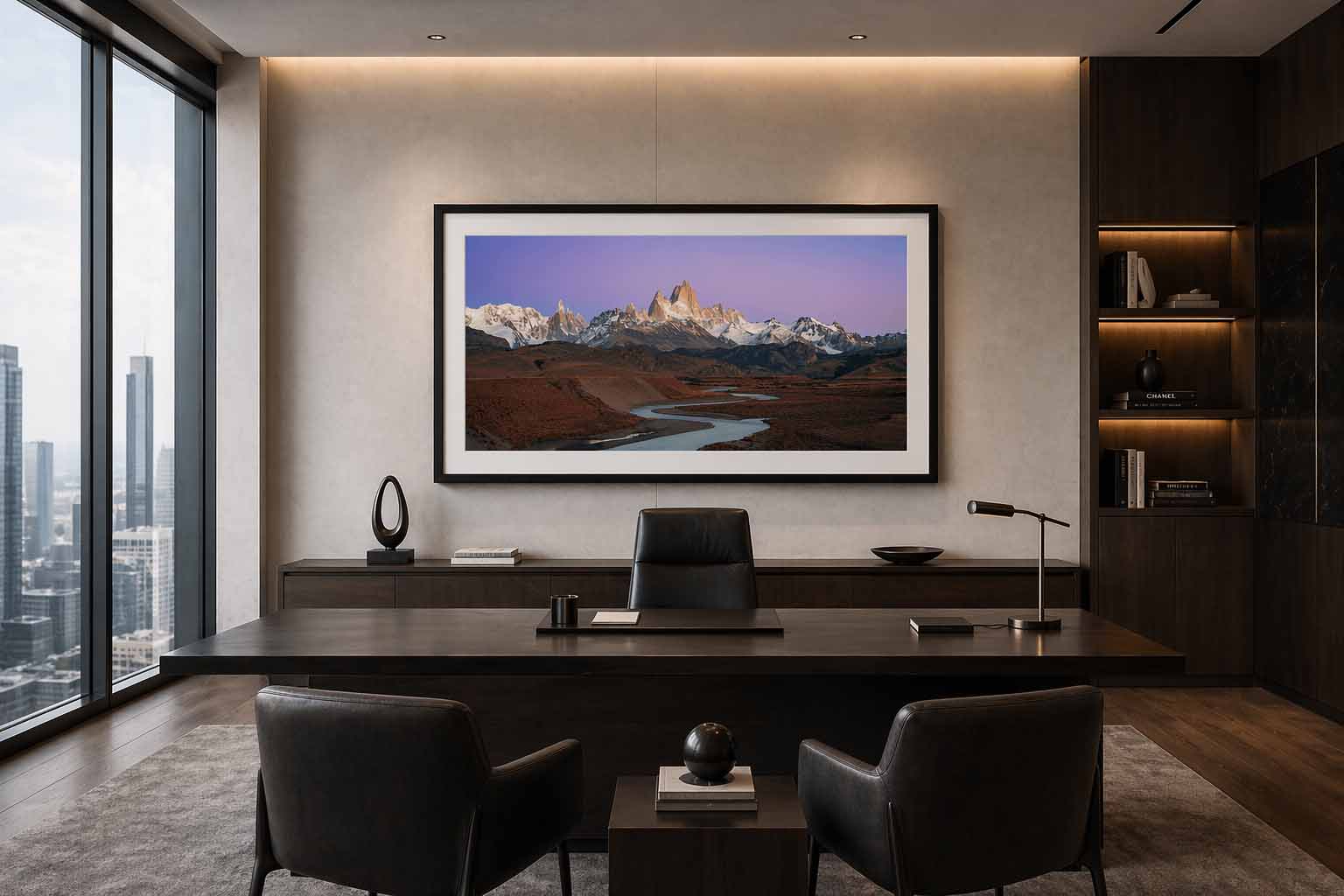 A large framed panoramic print of a Patagonian mountain range at sunrise hanging above a dark credenza in an executive office, with city windows to one side.
