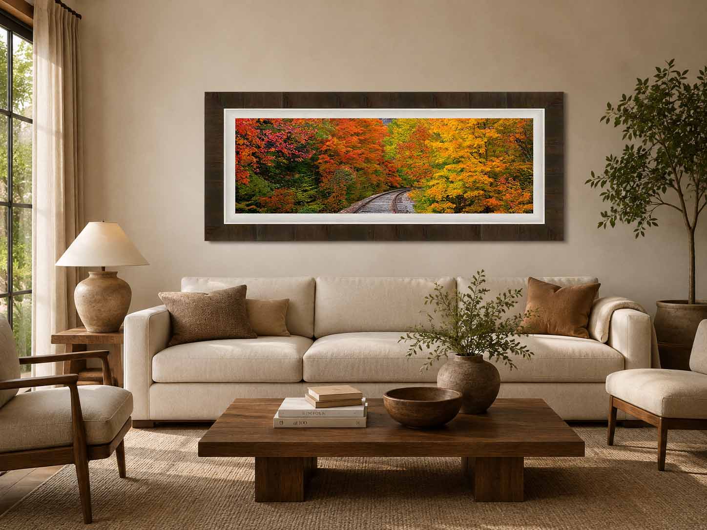 A panoramic photograph of a railroad curving through autumn maples, framed in dark ash with a white linen liner, hanging above a cream sofa in a warm neutral living room.