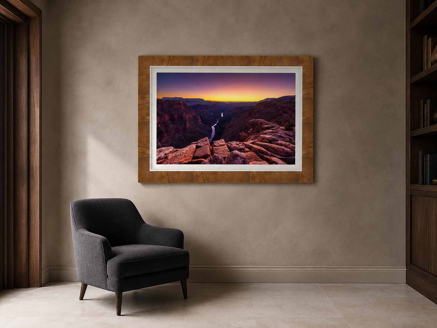 A photograph of the Colorado River from Toroweap Overlook at dusk, framed in warm brown wood with a white linen liner, above a charcoal reading chair in a plaster-walled study.