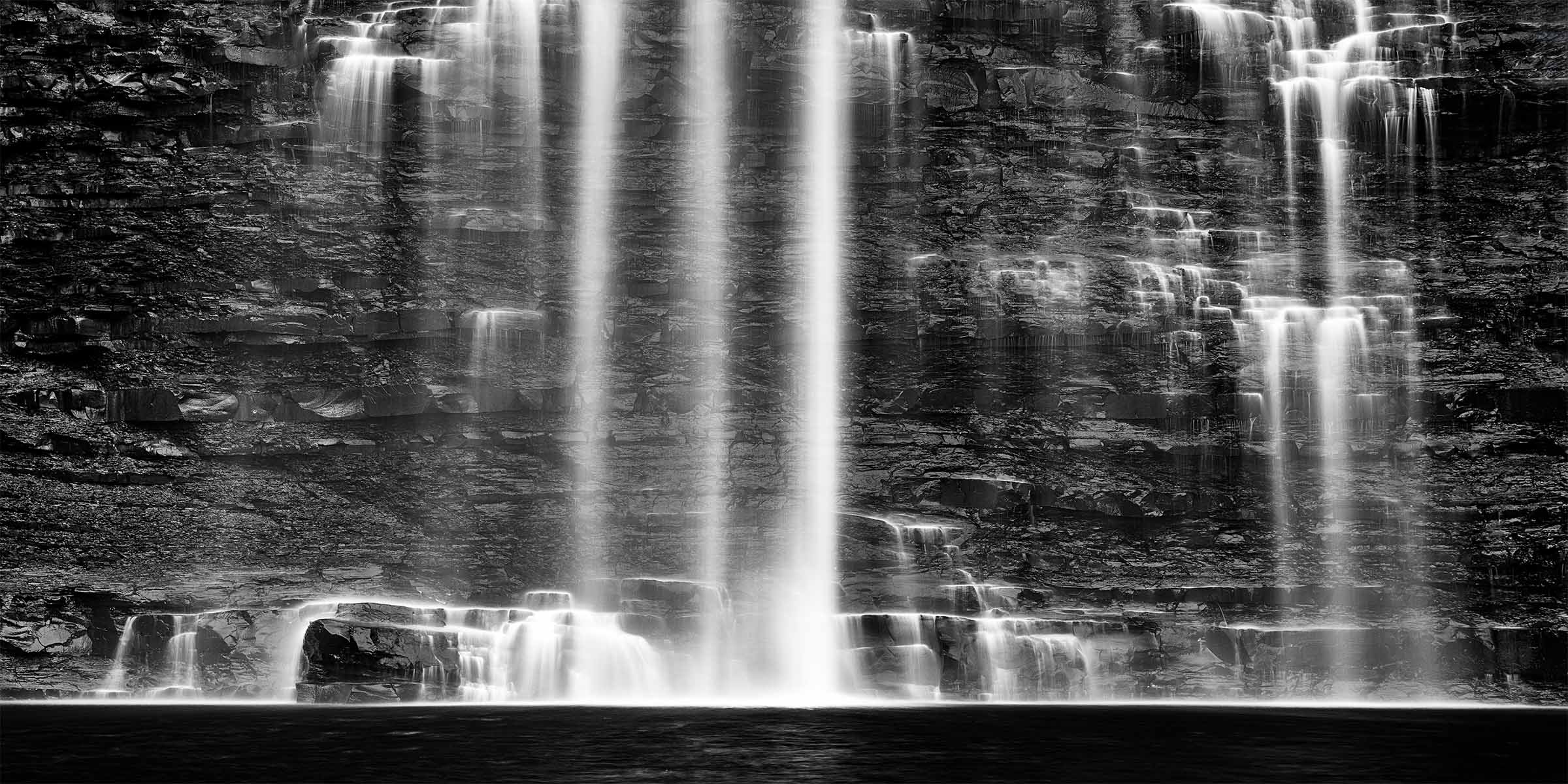 ARTIST&rsquo;S STORY Not all water falls the same way. At Watkins Glen in the Finger Lakes region of New York, the water does not plunge or crash or announce itself with the kind of force that demands your immediate attention. It descends. Slowly, deliberately, in multiple streams that find their own paths down the layered shale face, following the edges and contours of the rock with a gentleness that feels almost intentional. As if the water and the stone have reached an agreement over centuries about how this particular descent should be conducted. I moved in close on this day. The wider view was beautiful but it was the detail that held me, the way each stream of water caressed the rock edges as it passed, the way the layered shale caught and redirected the flow in ways that were never quite the same twice, the way the long exposure collapsed all of that movement into something between silk and light against the dark wet stone. The tighter crop was not a compositional choice so much as an honest one. This is what deserved to be seen. What I remember most is the sound. Not the roar of a powerful falls but something quieter and more complex, the layered conversation of multiple streams moving over rock at different speeds and volumes, filling the canyon with a natural acoustics that the mind receives not as noise but as relief. Research on the psychological effects of natural water sounds consistently finds that they reduce cortisol levels, lower heart rate, and produce measurable states of calm within minutes of exposure. Standing before the Weeping Wall I understood this not as data but as direct experience. There is something in the image of a wall that releases rather than holds that speaks to something most of us carry. The places in ourselves where we have been holding too long, where the accumulated weight of experience has been pressing against surfaces that were never designed to contain it indefinitely. The rock does not resist the water. It simply allows the release, slowly and continuously, along every available edge and surface, until the weight of what was held becomes the beauty of what flows. The Weeping Wall is a reminder that release is not weakness. It is what stone does when it has held long enough. It is what we do too, when we finally allow it. A  Limited Fine Art Edition Each image is released in limited editions, with edition size set by presentation and scale, so the most substantial works remain the most rare.