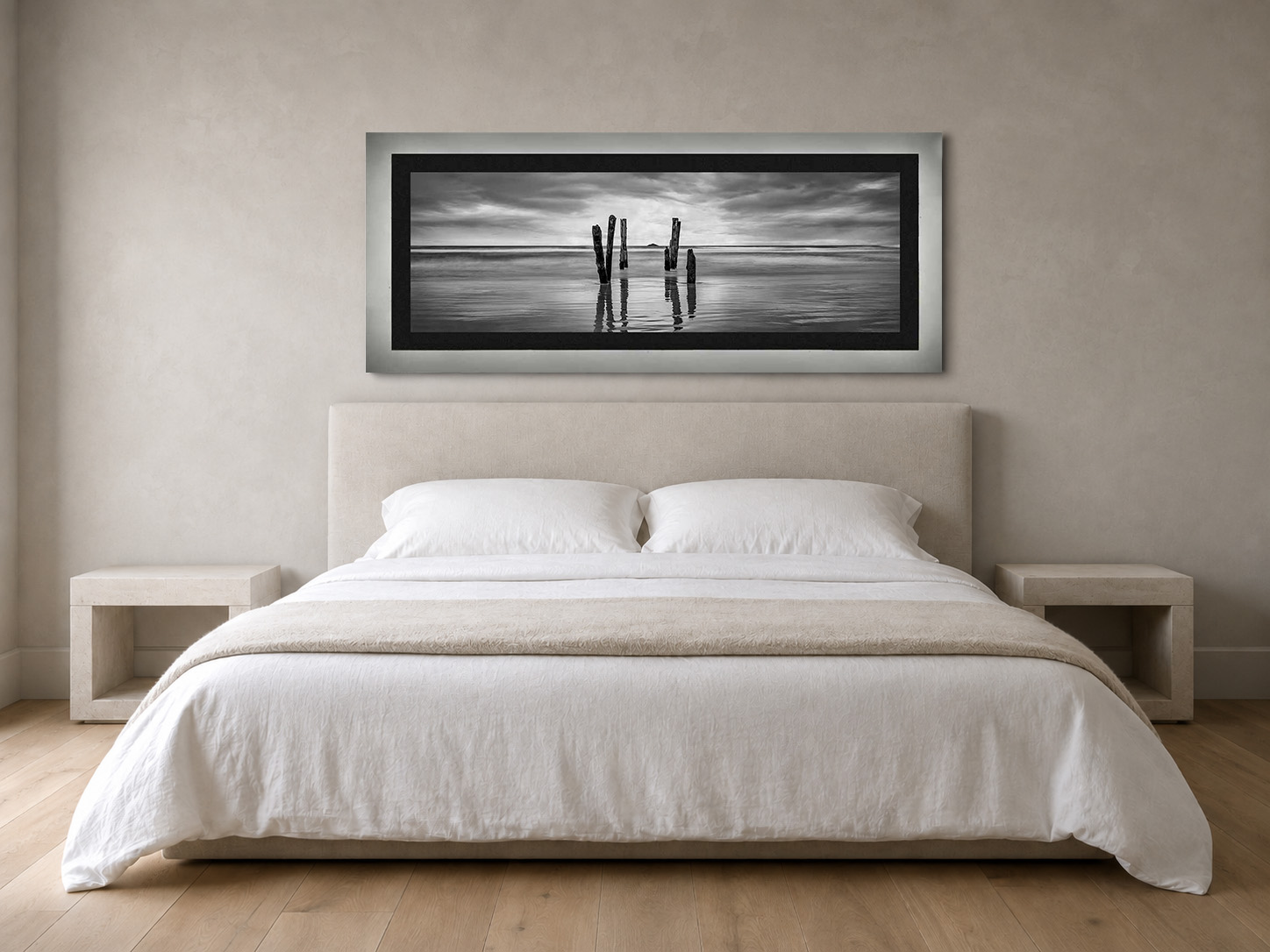 A black and white panoramic photograph of weathered beach poles, framed in pale gray with a black linen liner, hanging above a linen bed in a plaster-walled bedroom.