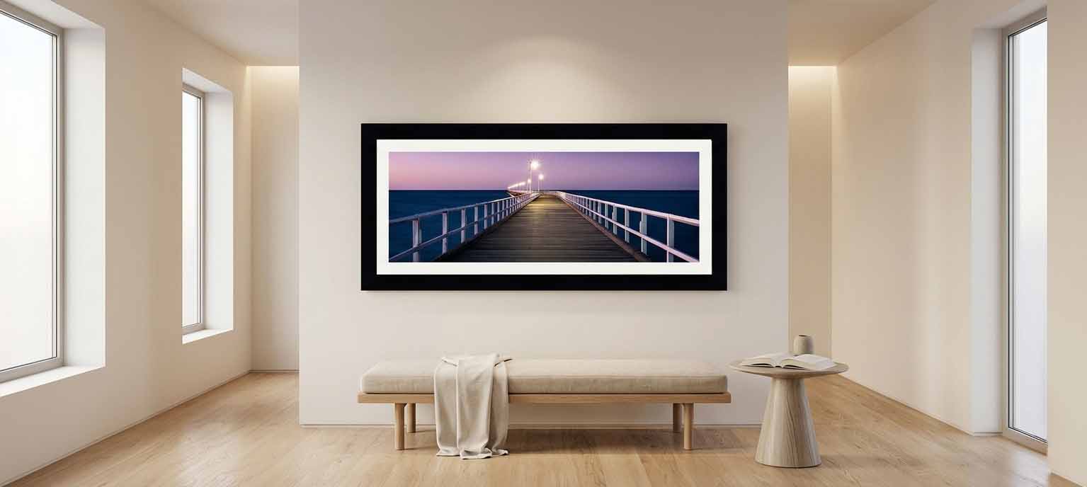 A large framed panoramic print of a lit pier at dusk hanging in a bright minimal entryway above a wooden bench with a folded throw and a small round side table