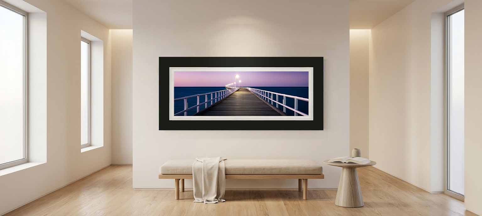 Urangan Pier at dusk with its lamps lit, framed in black with a white mat above a pale bench in a quiet sitting room.